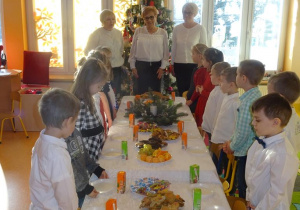 Pani dyrektor Maria Królikowska, pani Agnieszka Piekarska, pani Arleta Kalinowska oraz dzieci stoją wokół nakrytego białym obrusem i poczęstunkiem stołu.